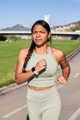 Focused african millennial woman jogging outdoors in park on sunny day, wearing sportswear and smartwatch. Expression shows energy, discipline and determination in active lifestyle.