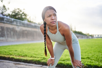 Exhausted african millennial woman resting after exercise in park, leaning forward with determination and focus. Expression shows effort, fatigue and resilience during outdoor fitness training.