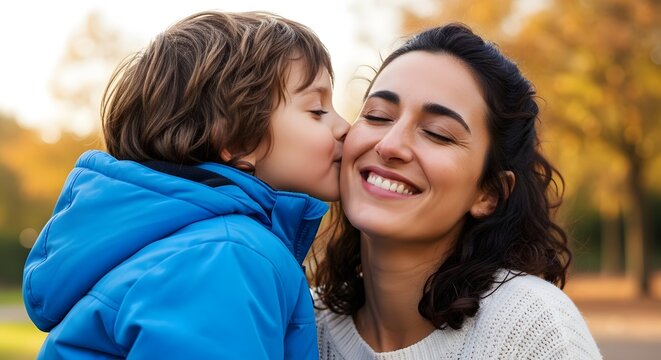 A young child kisses a smiling woman on the cheek. They're outside with autumn foliage in the background, showcasing a heartwarming familial moment.