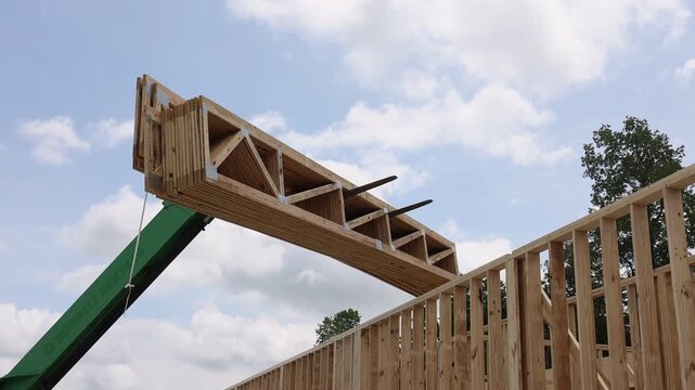 Telehandler crane carefully positions wooden rafters beams on construction site under bright blue sky.