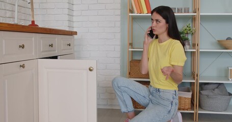 Homeowner checks kitchen sink cabinet, calling plumber at home. She reports a suspected clog or leak by phone and requests prompt repair service. Plumbing maintenance and household work concept