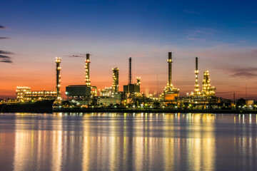 Fototapeta premium Illuminated oil refinery at dusk with colorful orange and blue sky. Petrochemical industrial complex with distillation columns, processing units, and bright lights reflecting on calm water surface.