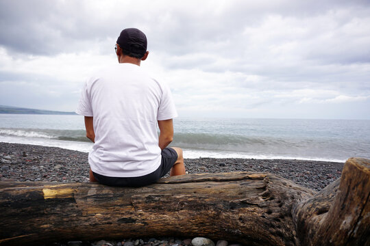 A man sitting alone on a long wooden log from behind on beach view background with cloudy sky. Deep in thought.