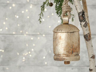 Background for personalized Christmas card. Metal vintage brass bell with rope and greenery against light plank wood backdrop and blurry twinkle fairy lights. Interior home Christmas decoration.