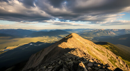 Golden hour light illuminates a rugged mountain ridge under a dramatic cloudy sky, with vast valleys stretching into the distance.