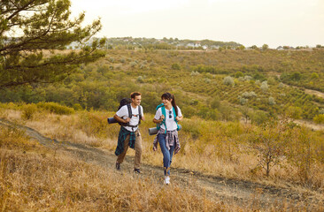 Obraz premium Hikers couple with backpacks on forest trail. They walk along a dry hillside, carrying mats and gear, enjoying travel and nature. Outdoor exploration, adventure on weekend, and hiking.
