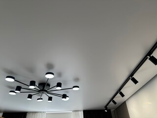 Stylish illuminated chandelier and track lighting on white ceiling in room