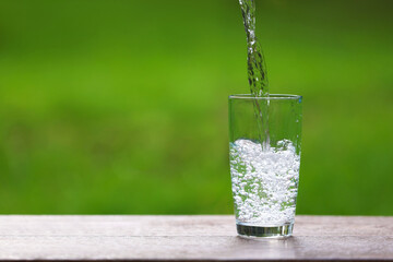 Pouring water into glass at wooden table against blurred green background, closeup. Space for text