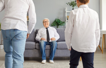 Obraz premium Joy of unity during family gathering. Happy elderly man sits comfortably on sofa and meets his adult son and grandson. Pensioner looks at man and boy walking towards him. Concept of family ties.