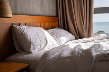 Unmade bed with white pillows and duvet in sunny hotel room with sea view