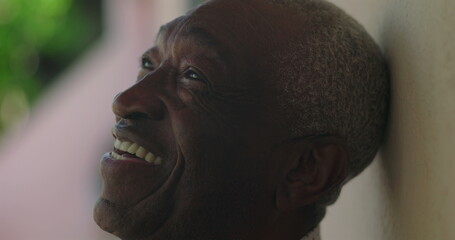 Elderly man of African descent smiling warmly, head tilted back, leaning against a wall, outdoors...