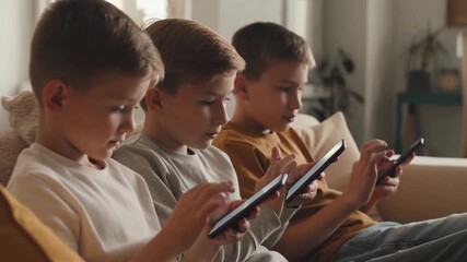 Three young boys are engrossed in using their devices, each focused on their respective screens. They are sitting together in a comfortable indoor setting. Stock Video - Powered by Adobe