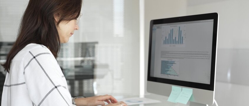 Businesswoman at glass office desk analyzing charts and graphs on computer screen, reviewing financial reports and performance data for planning. Business people concept
