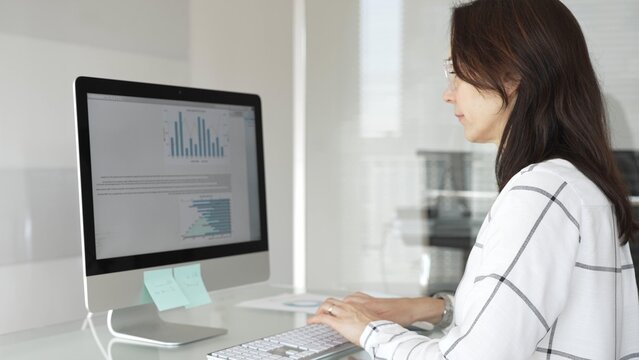 Businesswoman at glass office desk analyzing charts and graphs on computer screen, reviewing financial reports and performance data for planning. Business people concept - Powered by Adobe