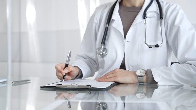 Female doctor in white coat with stethoscope writing patient notes on clipboard, completing medical records in clinic office. Medicine and health care