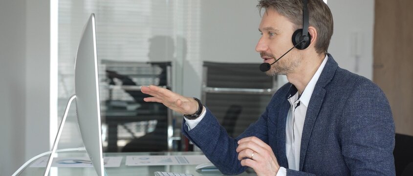 Male call center agent wearing a headset, providing online customer support and consulting clients during a video call. Business communication concept