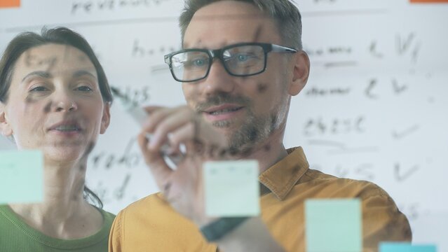 Professional team members brainstorming strategy, writing notes on transparent whiteboard with colorful sticky papers during collaborative meeting. Business people concept