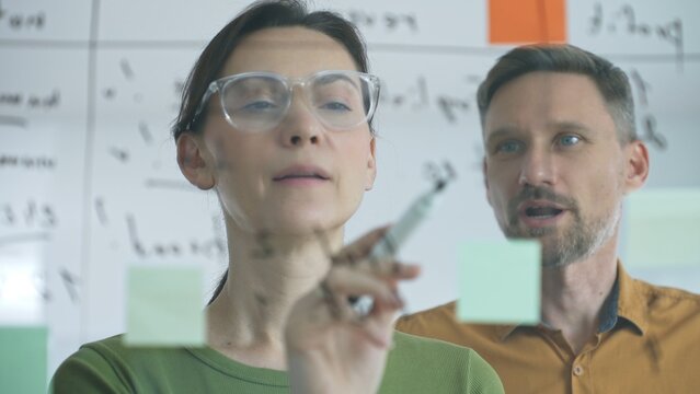 Businesswoman wearing glasses, writing on a glass board and organizing sticky notes, collaborates with adult businessman observing her strategy planning in a bright office
