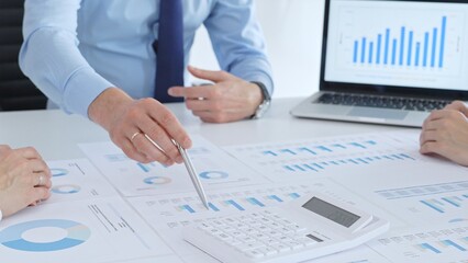 Business professional pointing at printed charts with a pen during a meeting, laptop showing bar graphs and calculator on table while team collaborates on financial analysis and planning