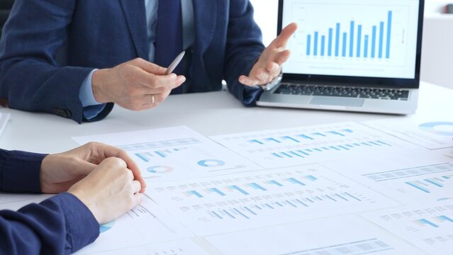 Business people reviewing financial charts and reports in office, using a laptop and calculator while engaging in discussions about strategy and performance