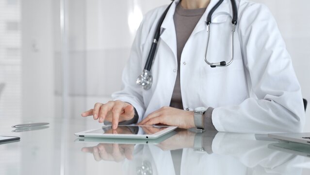 Physician in lab coat with stethoscope reviewing patient data on a tablet in a bright modern clinical office via touchscreen. Medicine and health care