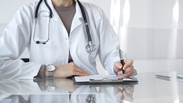 Female doctor in white coat with stethoscope writing patient notes on clipboard, completing medical records in clinic office. Medicine and health care