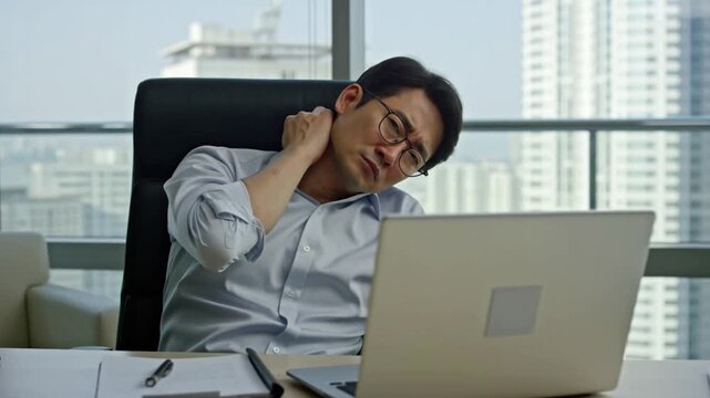 A stressed office worker is dealing with neck pain while working on a laptop. He is in a modern office with city views Stock Video