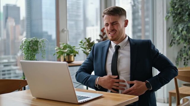 A businessman in distress clutching his stomach, possibly due to a health issue. The individual appears to be in pain, sitting at a desk with a laptop Stock Video