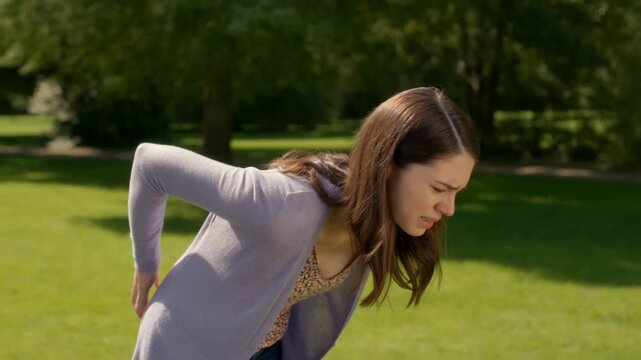A woman in a park bent over. She's dressed casually in a long-sleeved shirt and cardigan, and appears to be inspecting something on the ground. The background is a green lawn with trees. Stock Video