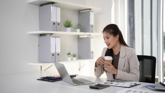 Asian professional woman works laptop office desk holding coffee cup business productivity virtual meeting professional workspace modern office environment