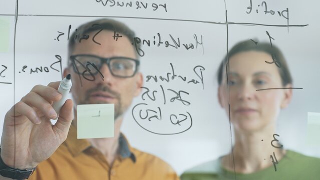 Business people brainstorming near glass board, using sticky notes for strategic planning, writing calculations, sharing innovative ideas during collaborative work session. close up - Powered by Adobe
