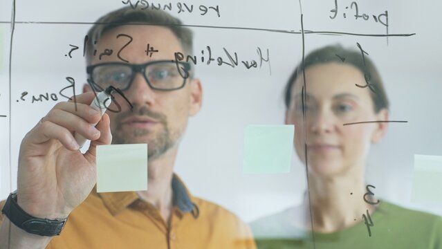 Business professionals brainstorming near glass board, using sticky notes for strategic planning, writing calculations, sharing innovative ideas during collaborative work session - Powered by Adobe