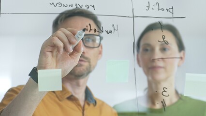 Professional team members brainstorming strategy, writing notes on transparent whiteboard with colorful sticky papers during collaborative meeting in office. Business people at work