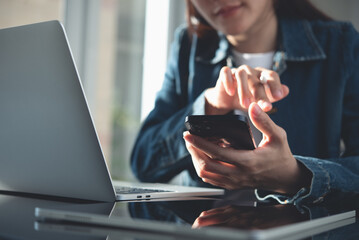 Close up, young asian woman using mobile phone while working on laptop computer in office. Casual business woman, freelancer online working at home office