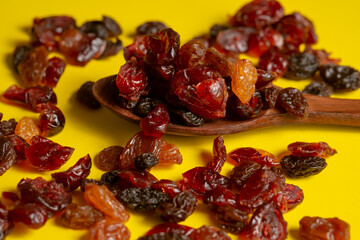 A macro close-up of a dark wooden spoon filled with a mixture of dark raisins (dried grapes) and bright red, translucent dried cranberries. The fruits are scattered on a contrasting, vibrant yellow ba