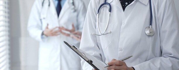 A medical professional with a stethoscope writes on a clipboard while a doctor and female assistant collaborate in a clinic, discussing patient data and healthcare information