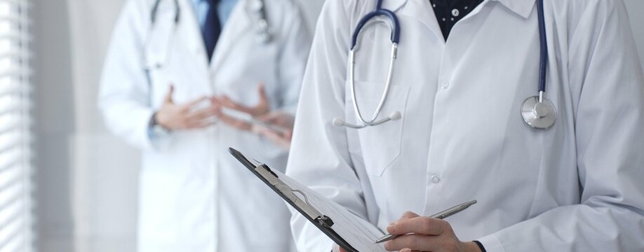 A medical professional with a stethoscope writes on a clipboard while a doctor and female assistant collaborate in a clinic, discussing patient data and healthcare information