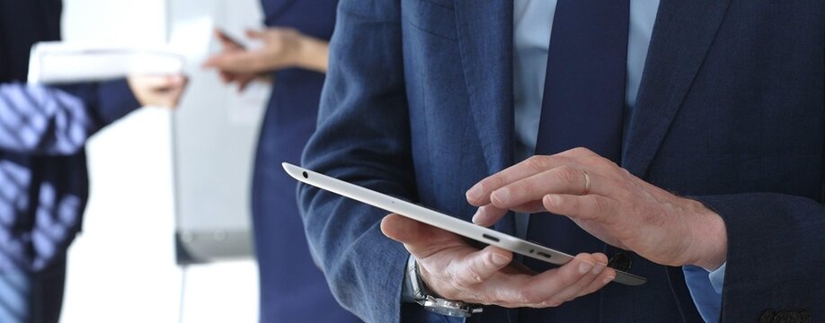Businessman in a blue suit using a tablet during a corporate meeting, interacting with the touchscreen as blurred colleagues collaborate in the background, in office. Business people concept