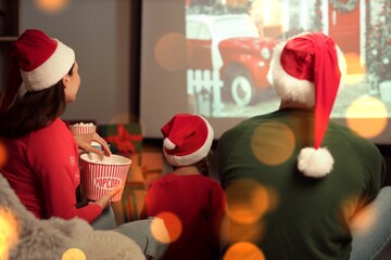 Little girl and her parents in Santa hats and with popcorn watching Christmas movie on projector...