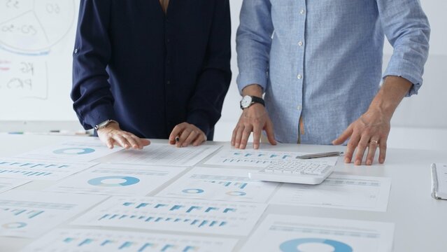 Focused business professionals analyzing financial charts and graphs, discussing market trends and investment strategies while leaning over white desk surface.