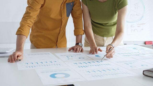 Two business professionals are examining financial data and reports, pointing at charts and graphs displayed on a table during a collaborative office meeting