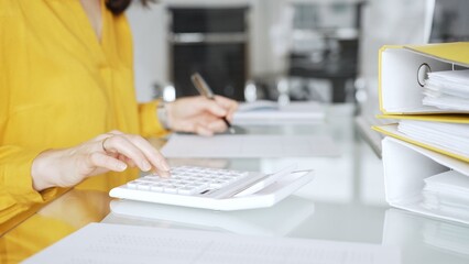 Woman in yellow blouse calculating finances at office desk, using calculator and writing,...