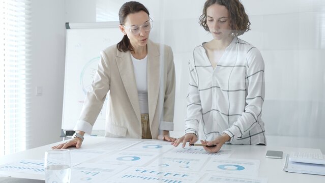 Professional businesswomen analyzing financial charts, collaborating in sleek corporate workspace, highlighting data. Business people concept