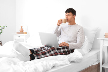 Young man with laptop drinking coffee in bedroom