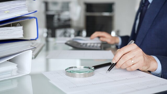 Business person inspecting financial documents with magnifying glass, calculating data for audit and analysis. Audit and taxes in business