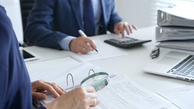 Business colleagues are working together, analyzing financial documents with a magnifying glass, calculating and taking notes in the office. Audit and taxes in business