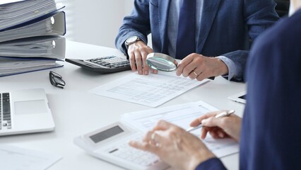Close up of male financial analysts with female colleague examining documents, using magnifying glass and calculator at workplace in office. Audit and taxes in business