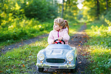 Little preschool girl driving big vintage toy car. Happy child having fun with playing outdoors. Active preschooler child enjoying warm autumn day in forest. Smiling stunning kid playing