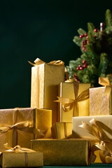 Golden Christmas presents with ribbons beside pine tree on dark green background, festive still life for winter holiday celebration scene.