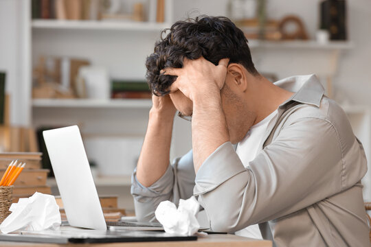 Stressed male screenwriter with laptop writing script at table in office